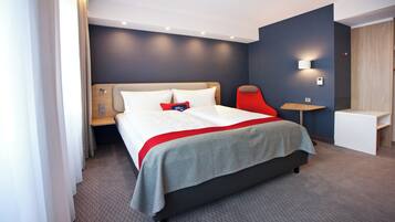 Una habitación de hotel moderna con una cama grande, una mesita de noche, un escritorio pequeño y una lámpara de pared.