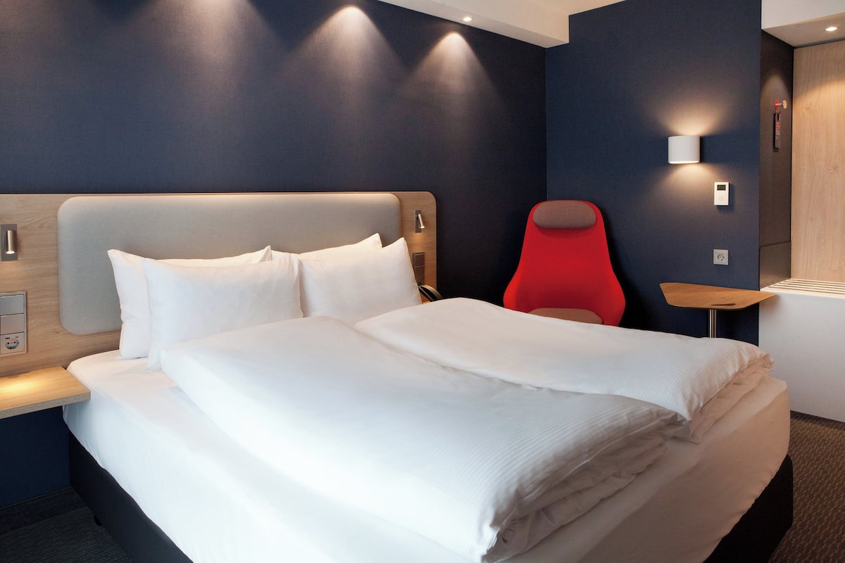 Een moderne hotelkamer met een groot bed, een rode stoel en een klein tafeltje met een lamp.