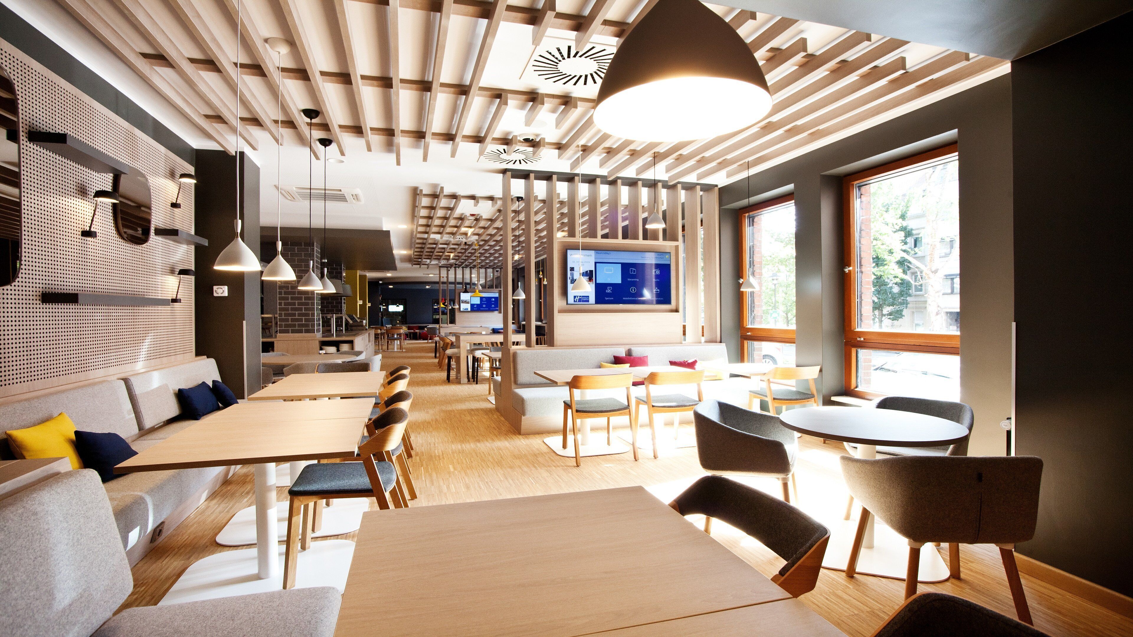 Un comedor moderno con mesas y sillas de madera, una amplia pantalla de televisión y un techo con listones de madera e iluminación colgante.