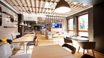 A modern dining area with wooden tables and chairs, a large TV screen, and a ceiling with wooden slats and pendant lighting.