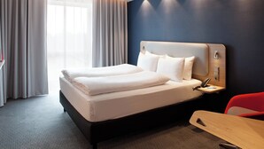 Una habitación de hotel moderna con una cama grande, una mesita de noche con lámpara y una silla roja.