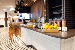Een modern hotel ontvangstgebied met een balie, barstoelen en een presentatie van fruit en snacks.