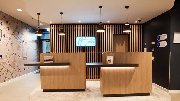 Zona de recepción con mostrador de madera, dos espacios de asiento y una pantalla grande con el logotipo de Holiday Inn Express.