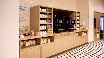Une chambre d’hôtel équipée d’un coin cuisine, comprenant une machine à café, des étagères avec des tasses et un panneau indiquant la disponibilité des boissons.