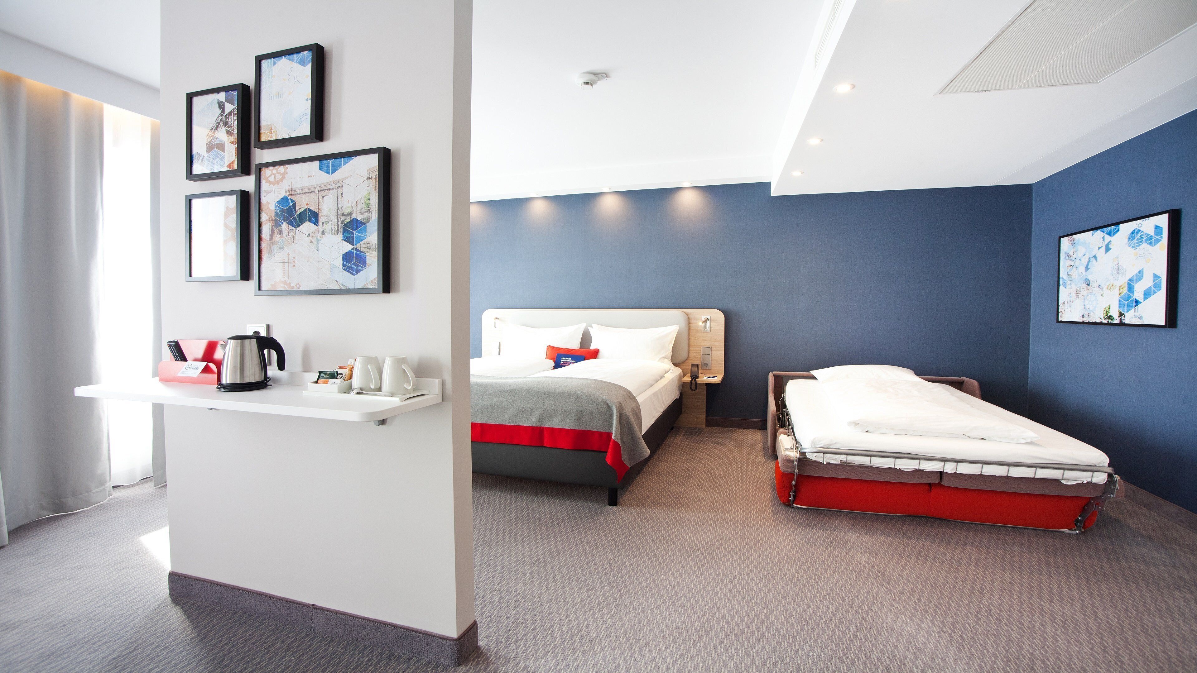 A hotel room with a bed, bedside table, and a desk with a kettle and coffee cups.