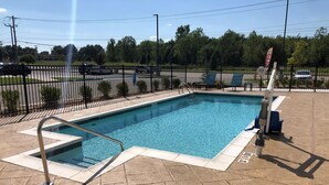 Une piscine rectangulaire avec un escalier en métal et un panneau de sécurité, entourée d’une terrasse dallée, elle-même bordée d’une clôture en métal et d’un mât portant un drapeau.