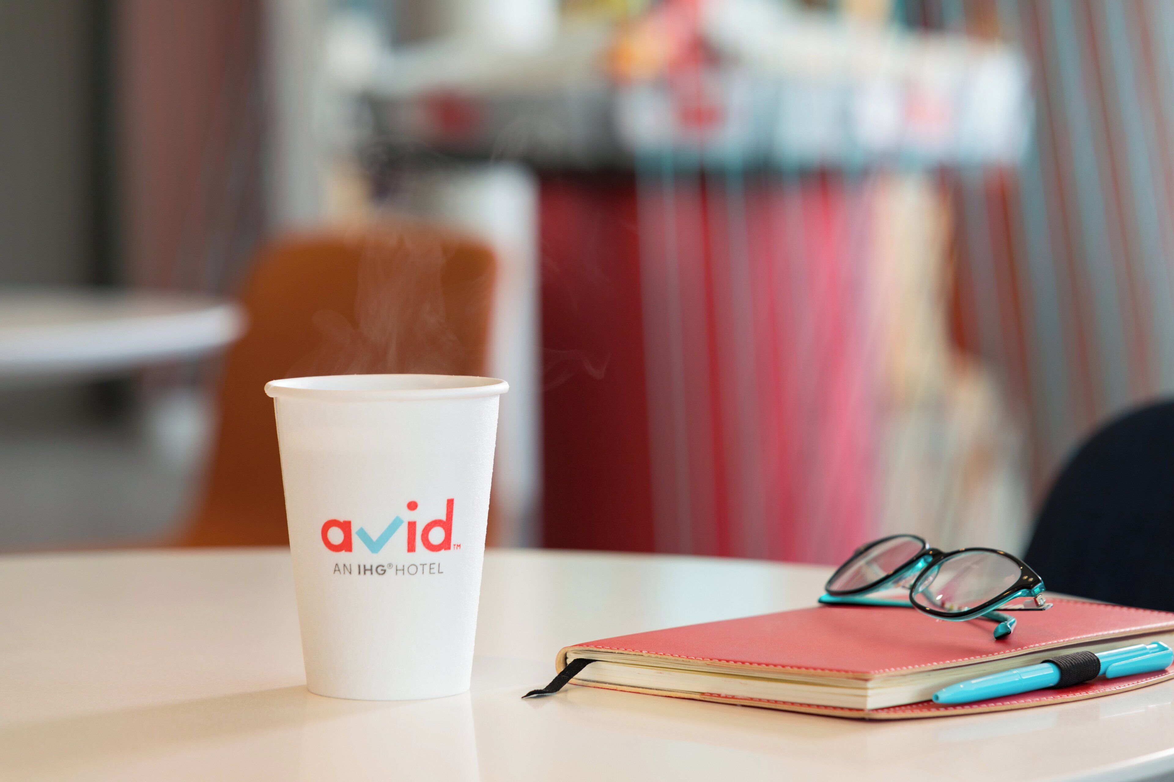 A white cup with 'avid' logo, a red notebook, and a pen on a table.
