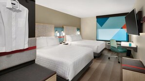 Une chambre d’hôtel avec deux lits, un bureau, une chaise, une télévision et une fenêtre donnant sur un paysage aux formes géométriques.