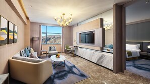 A modern hotel room with a large TV, comfortable seating, a small table, and a view of the mountains.