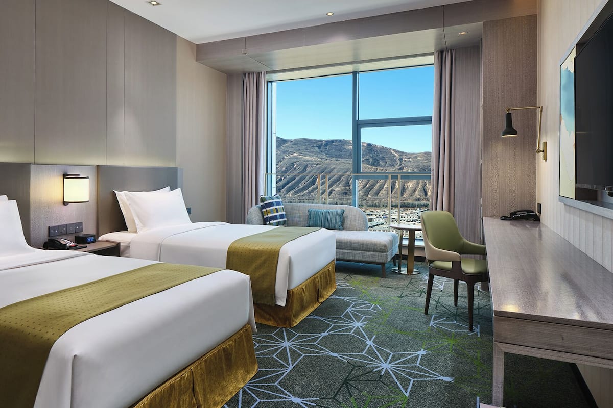 Ein Hotelzimmer mit zwei Betten, einem Schreibtisch, einem Stuhl, einem Fernseher und einem großen Fenster mit Blick auf die Berge.