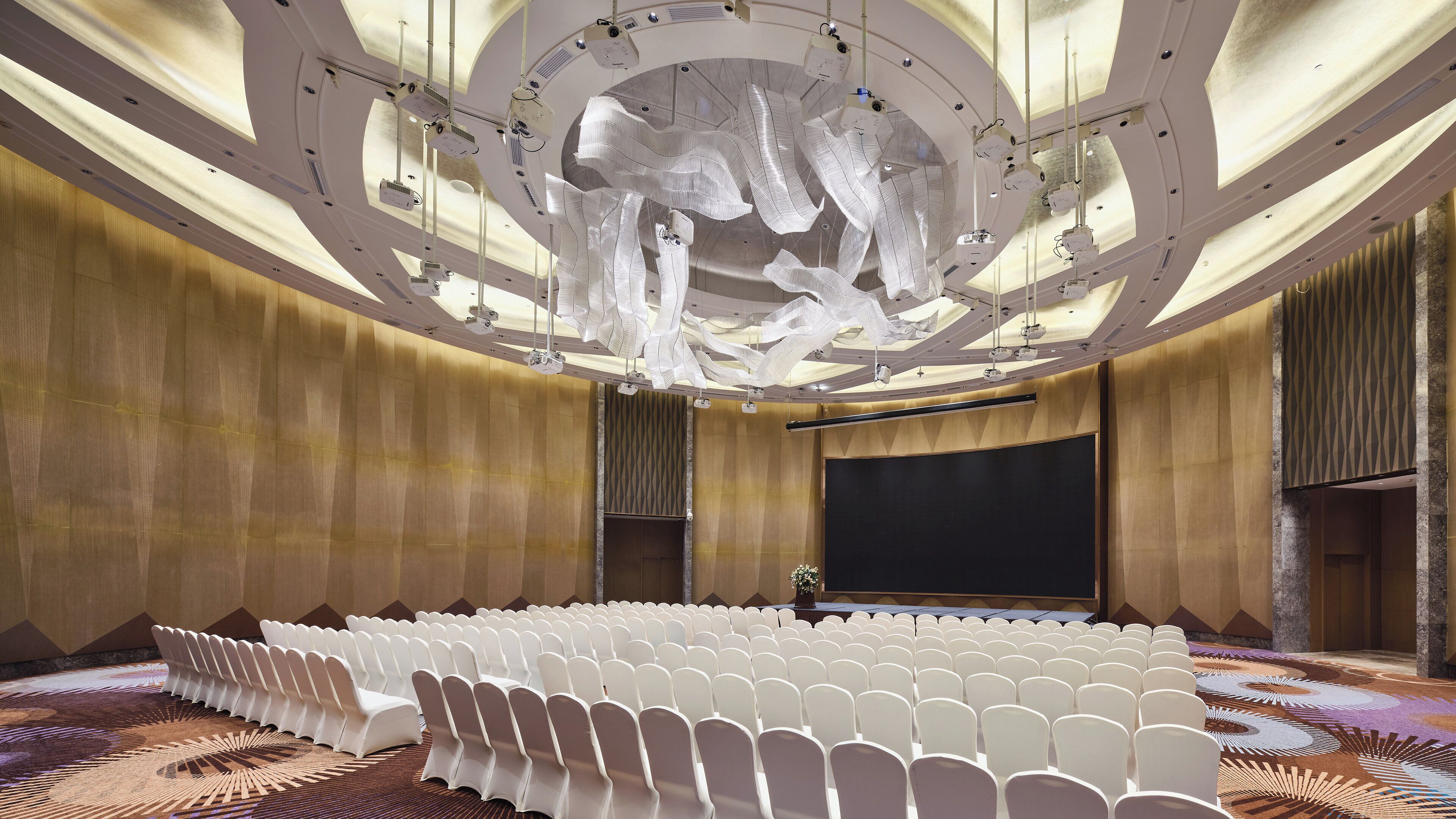 A conference room with rows of white chairs, a large screen, and a ceiling with hanging projectors.