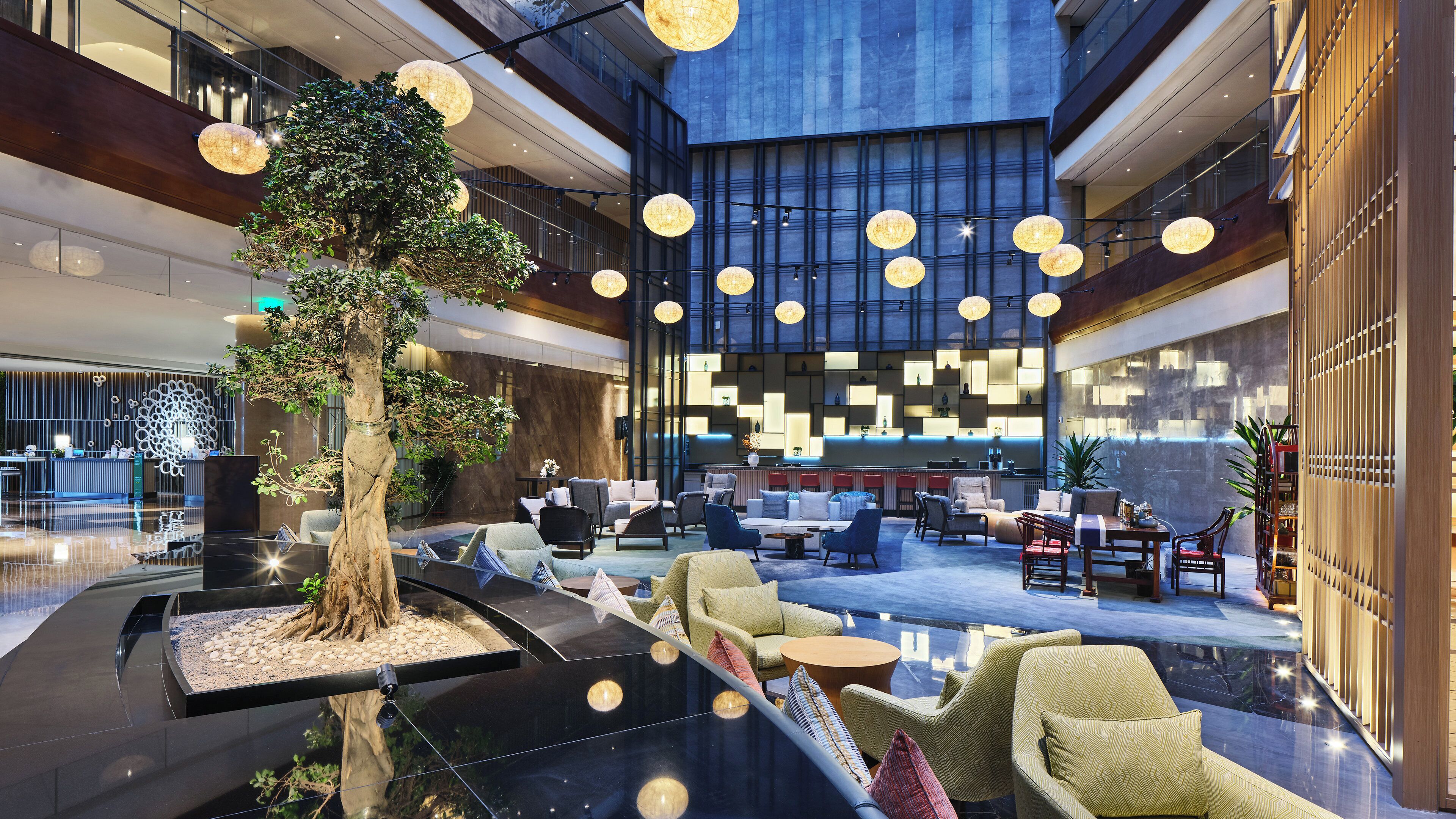 Un lobby amplio con un árbol central, zonas de estar y un mostrador de recepción moderno.