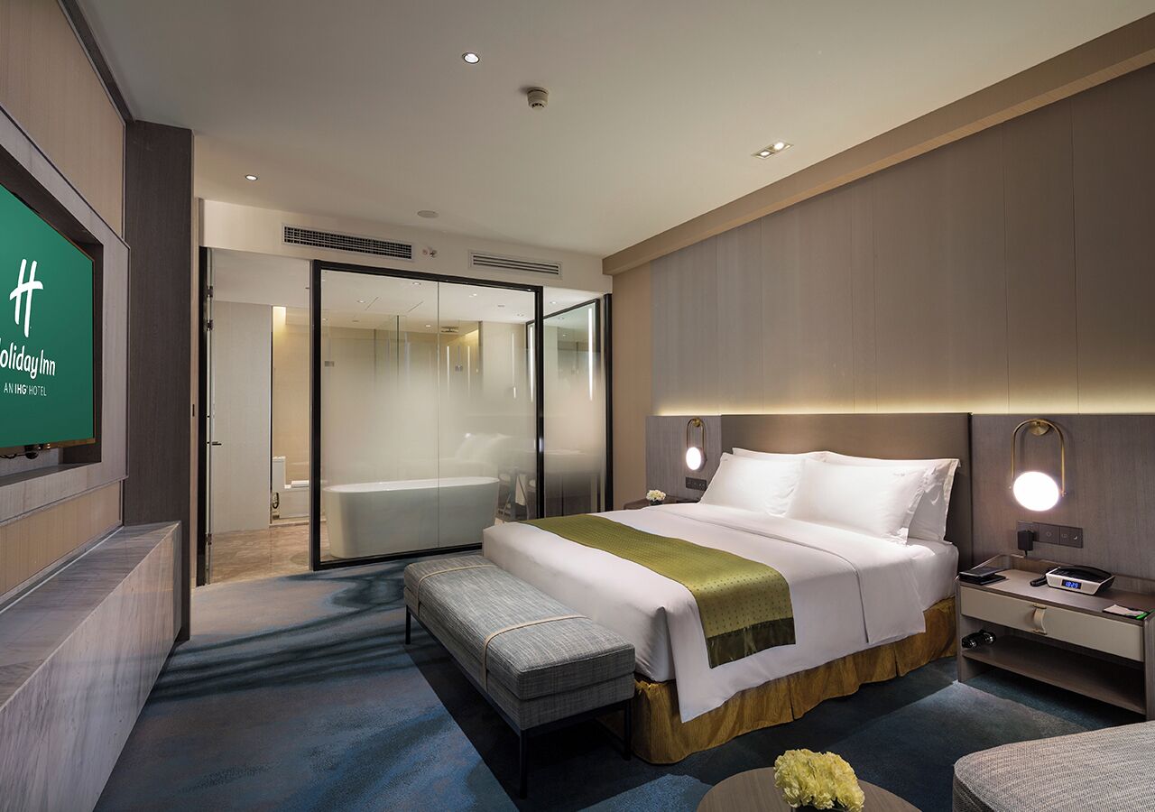 A modern hotel room with a large bed, a bench, a bedside table with a lamp, and a bathroom visible through a glass door.