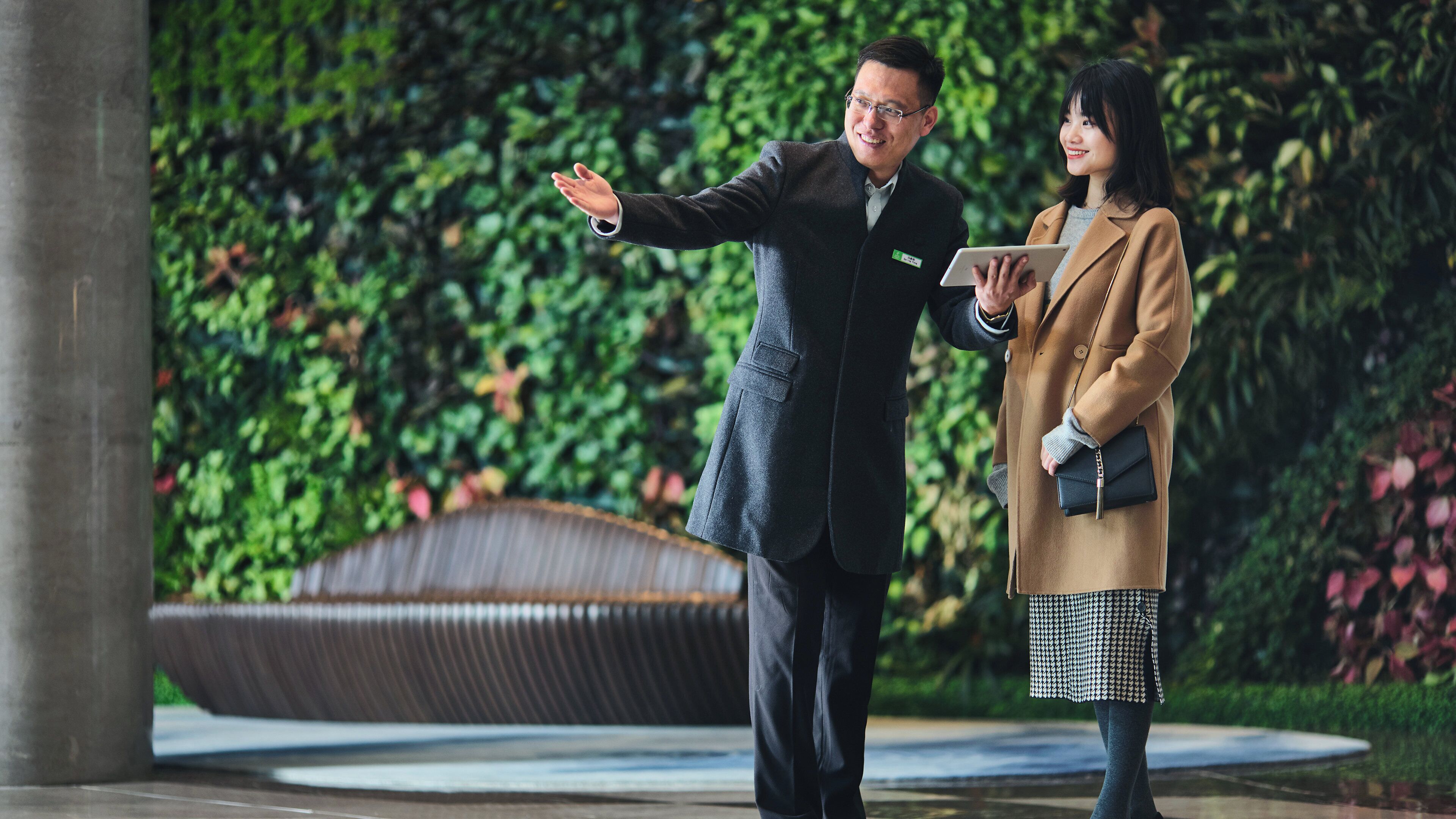 Two individuals standing in an outdoor area with a backdrop of greenery and a curved structure.