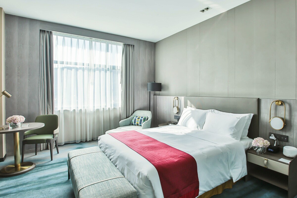 A hotel room with a bed, a desk, a chair, a nightstand, and a window with curtains.