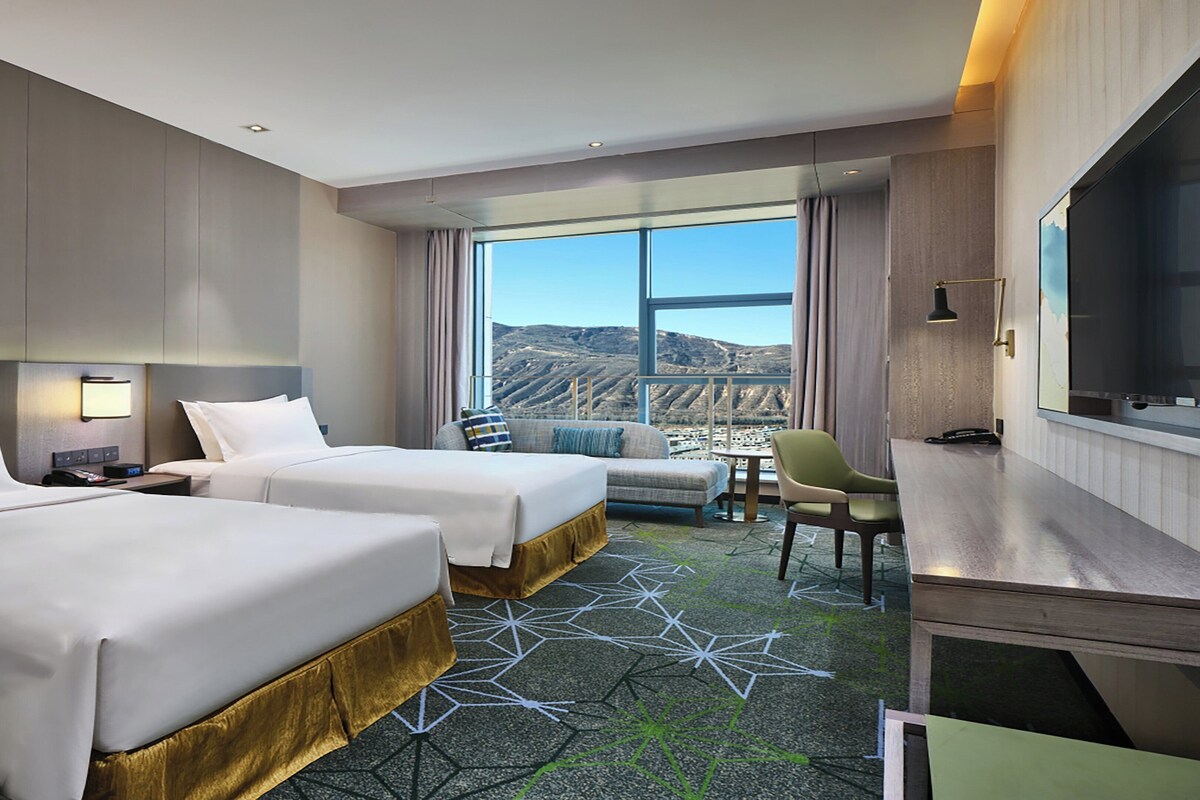A hotel room with two beds, a desk, a chair, and a large window with a view of the mountains.