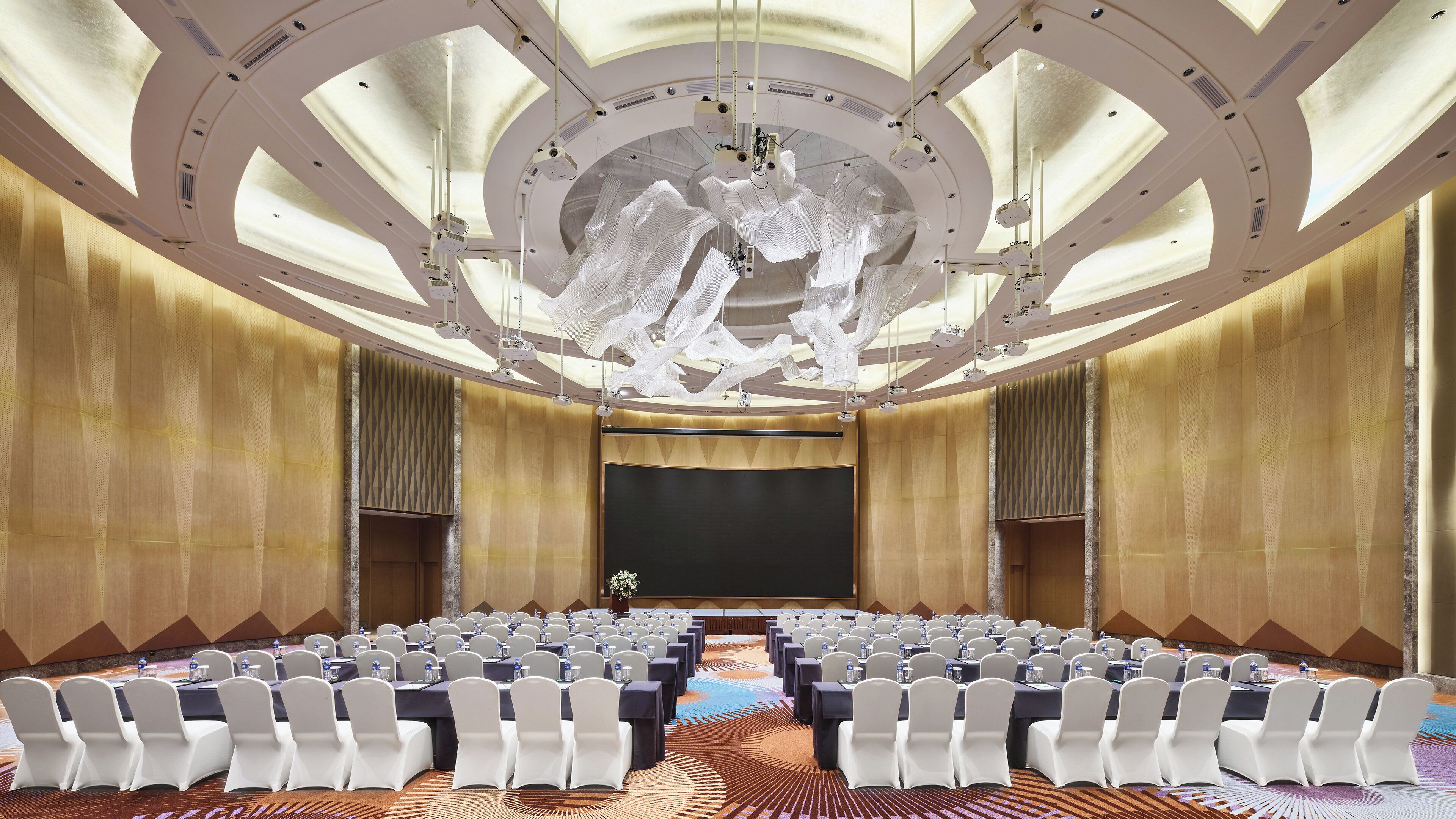 Un salón de conferencias con filas de sillas blancas, un escenario con una pantalla grande y un techo con una lámpara de diseño.