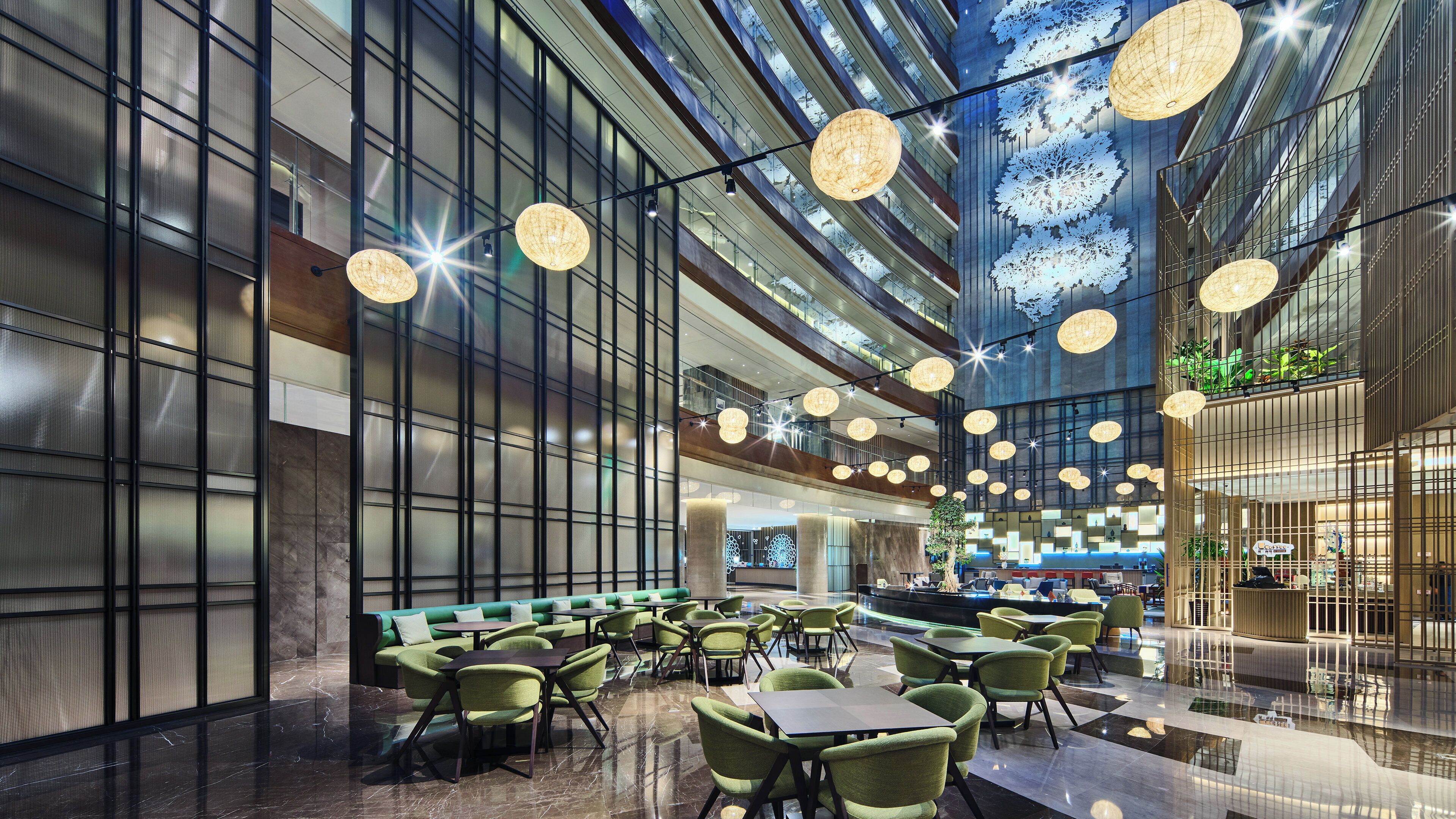 A modern interior with a high ceiling, hanging spherical lights, and a seating area with green upholstered chairs and tables.