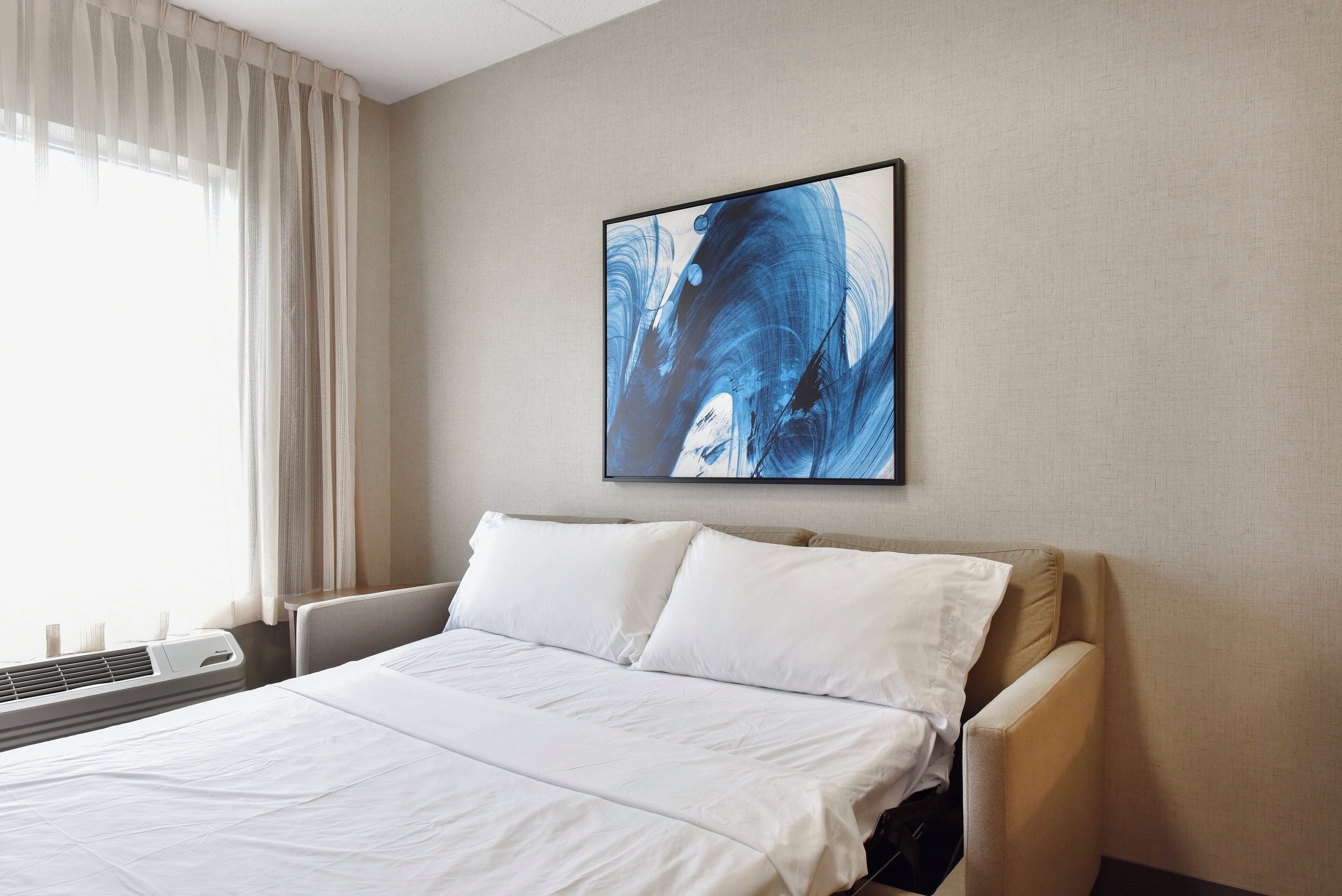 A hotel room with a bed, a window with curtains, and a painting on the wall.