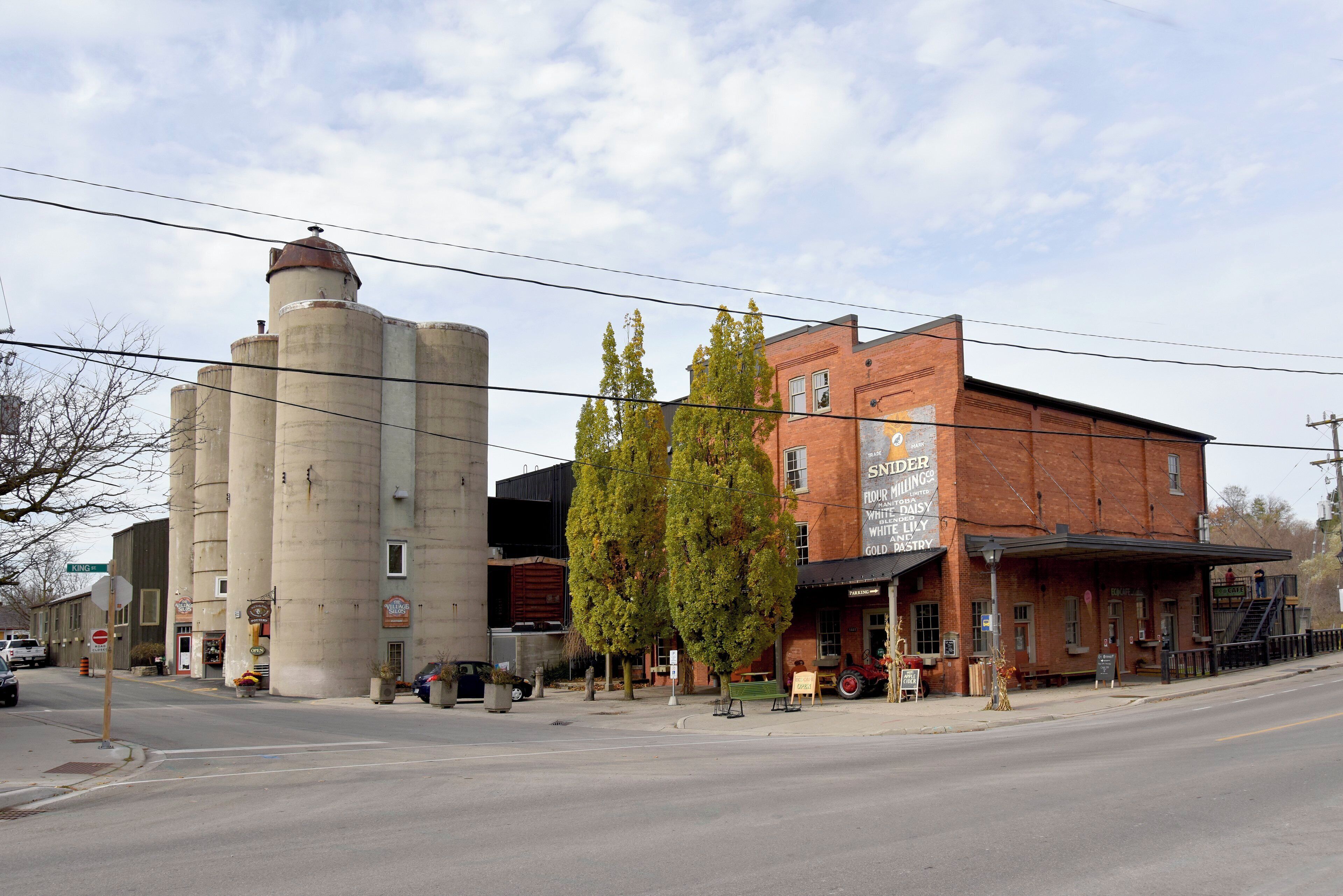 A street view with a grain silo, a red brick building with a sign for a flour mill, and a tree in the foreground.