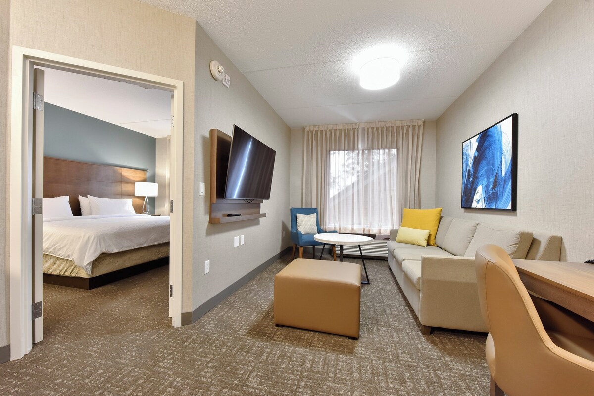 A hotel room with a bed, a sofa, a coffee table, a TV, and a painting on the wall.
