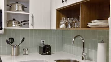 Une cuisine avec des meubles blancs, un évier en acier inoxydable, un grille-pain et une étagère en bois où sont rangés de la verrerie et des assiettes.