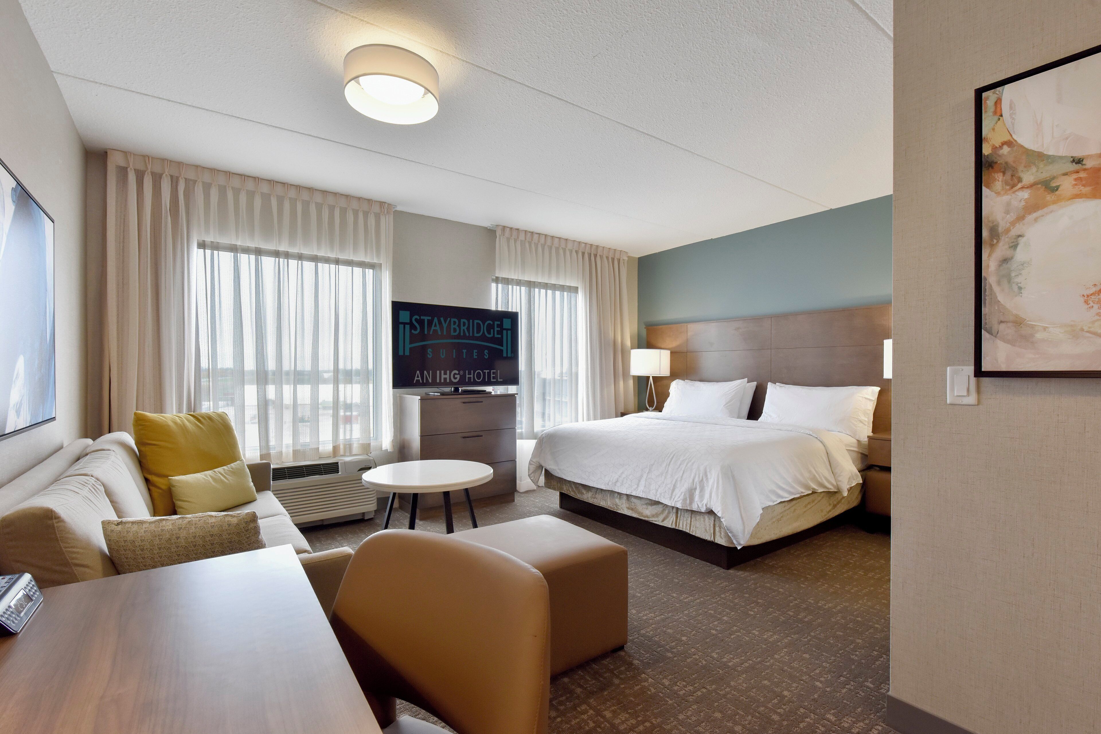 Une chambre d’hôtel avec un lit, un canapé, une petite table, une télévision et un panneau indiquant « Staybridge Suites ».