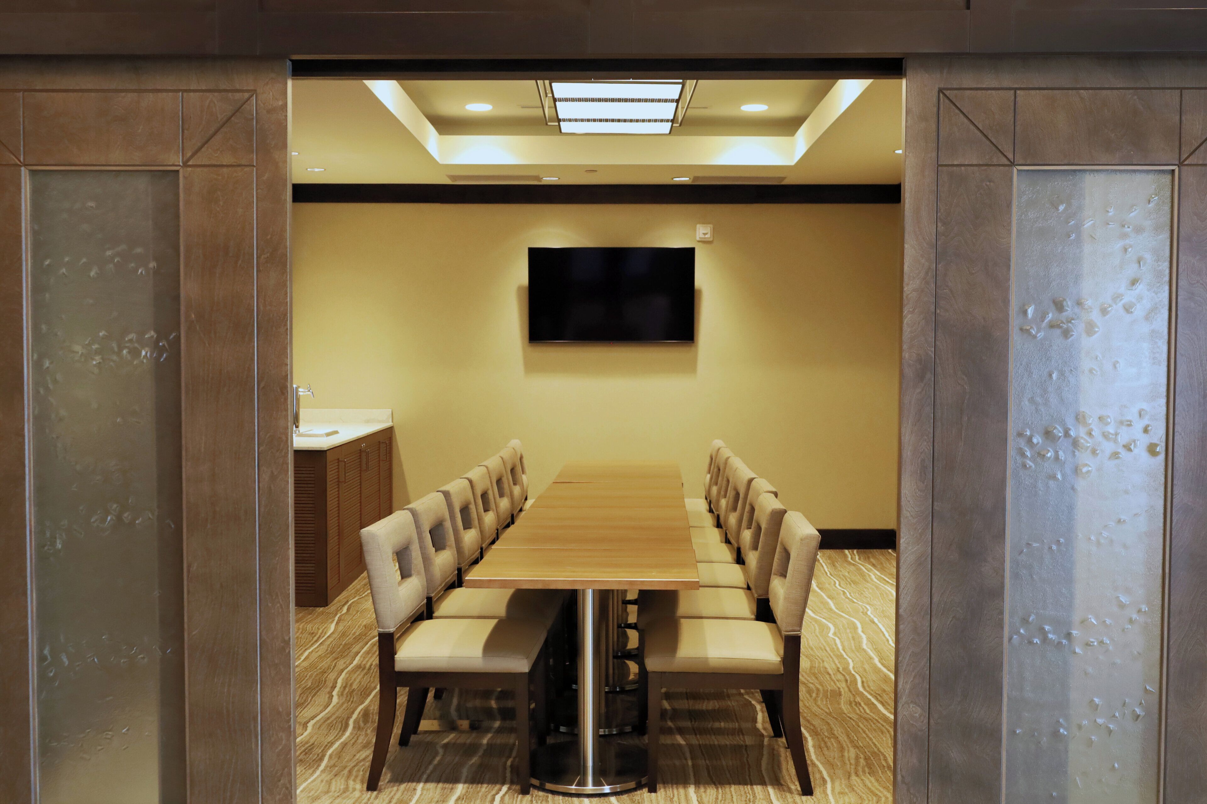 A conference room with a long table, chairs, a television, and a small kitchenette.