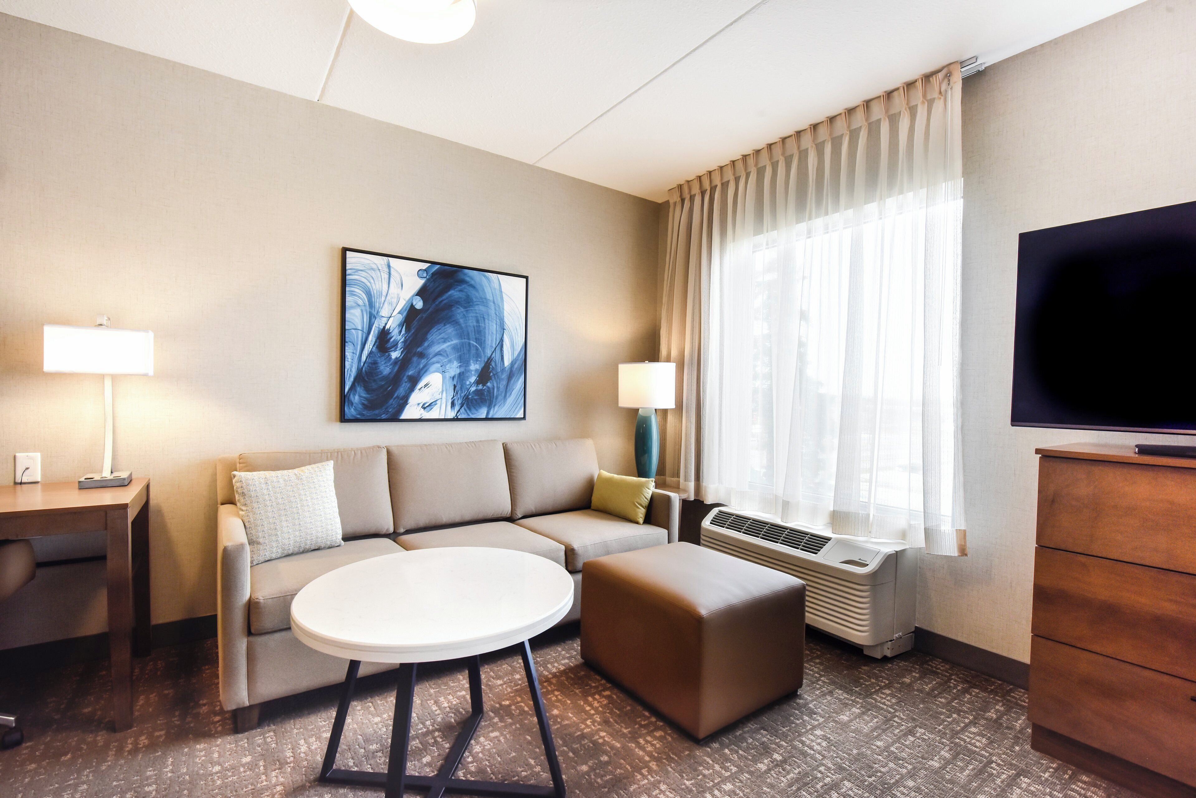 A hotel room with a sofa, ottoman, round table, TV, air conditioning unit, and a painting on the wall.