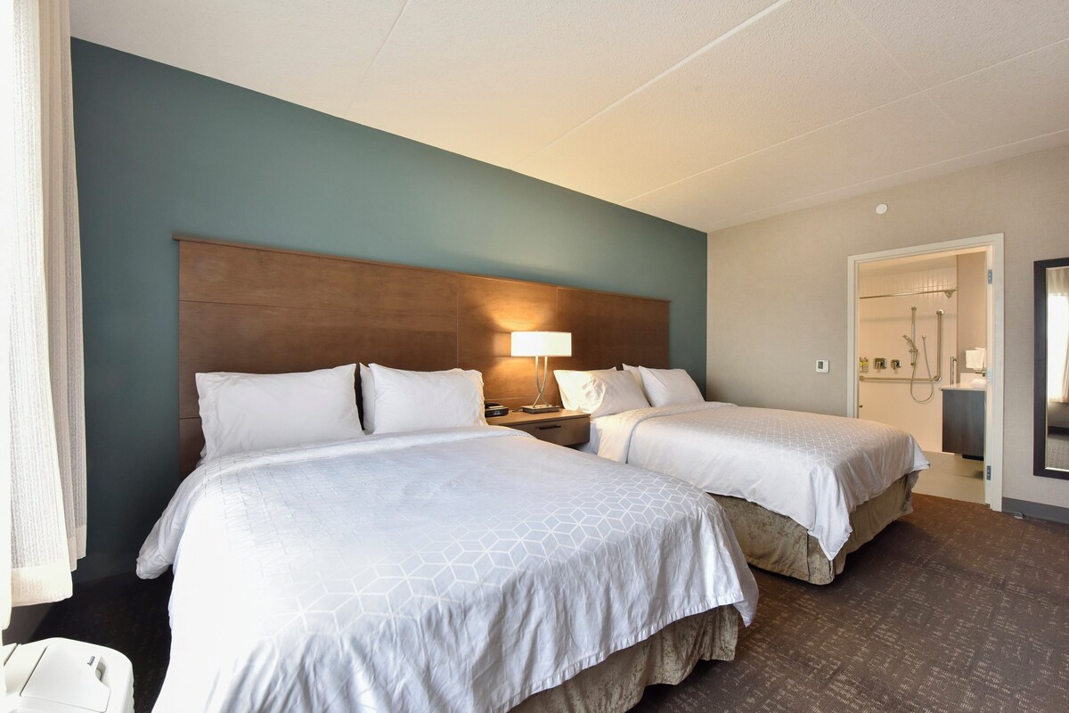 A hotel room with two beds, a nightstand, a lamp, a bathroom with a shower, and a mirror.