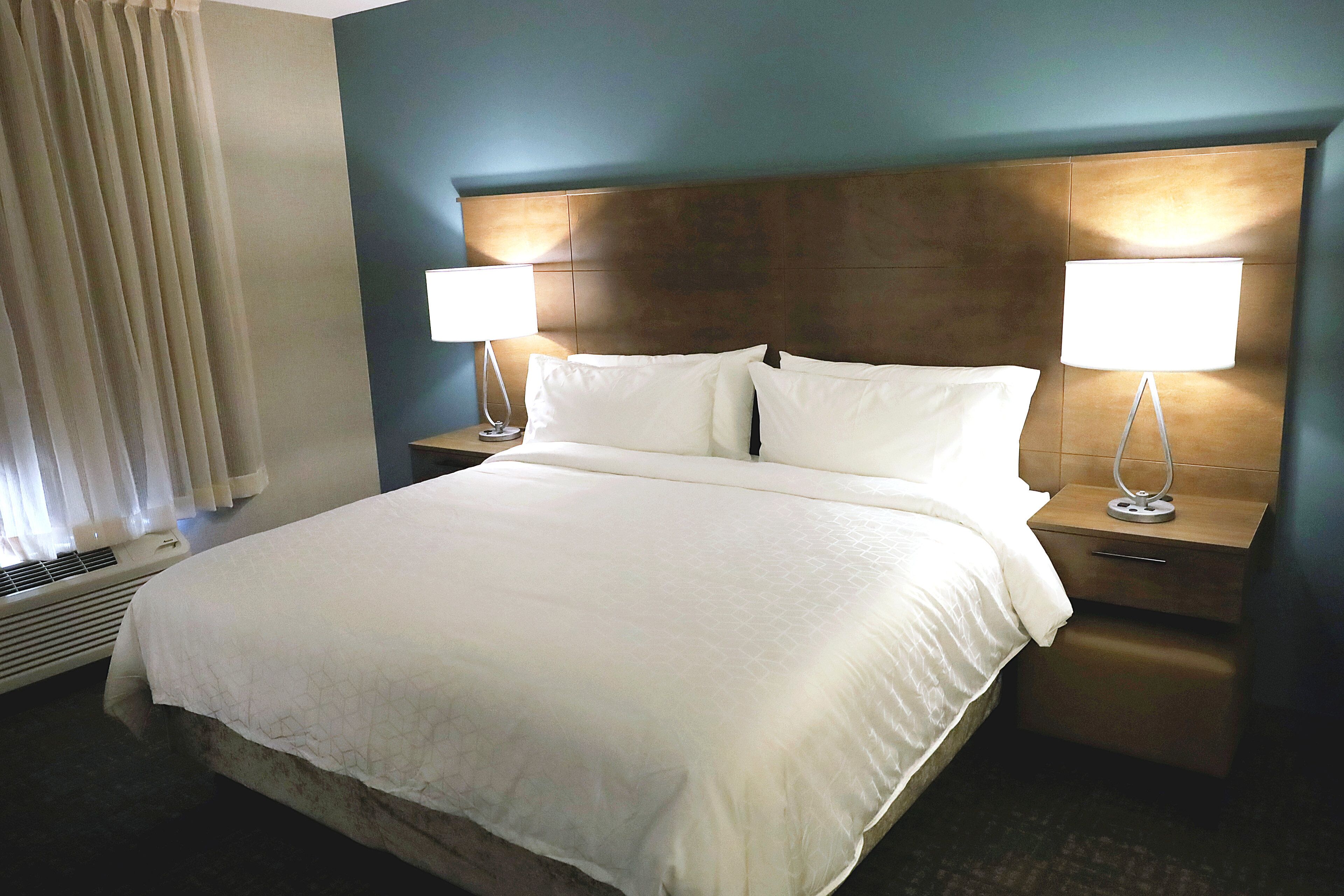 A hotel room with a large bed, two bedside tables with lamps, a window with curtains, and a wall-mounted heater.