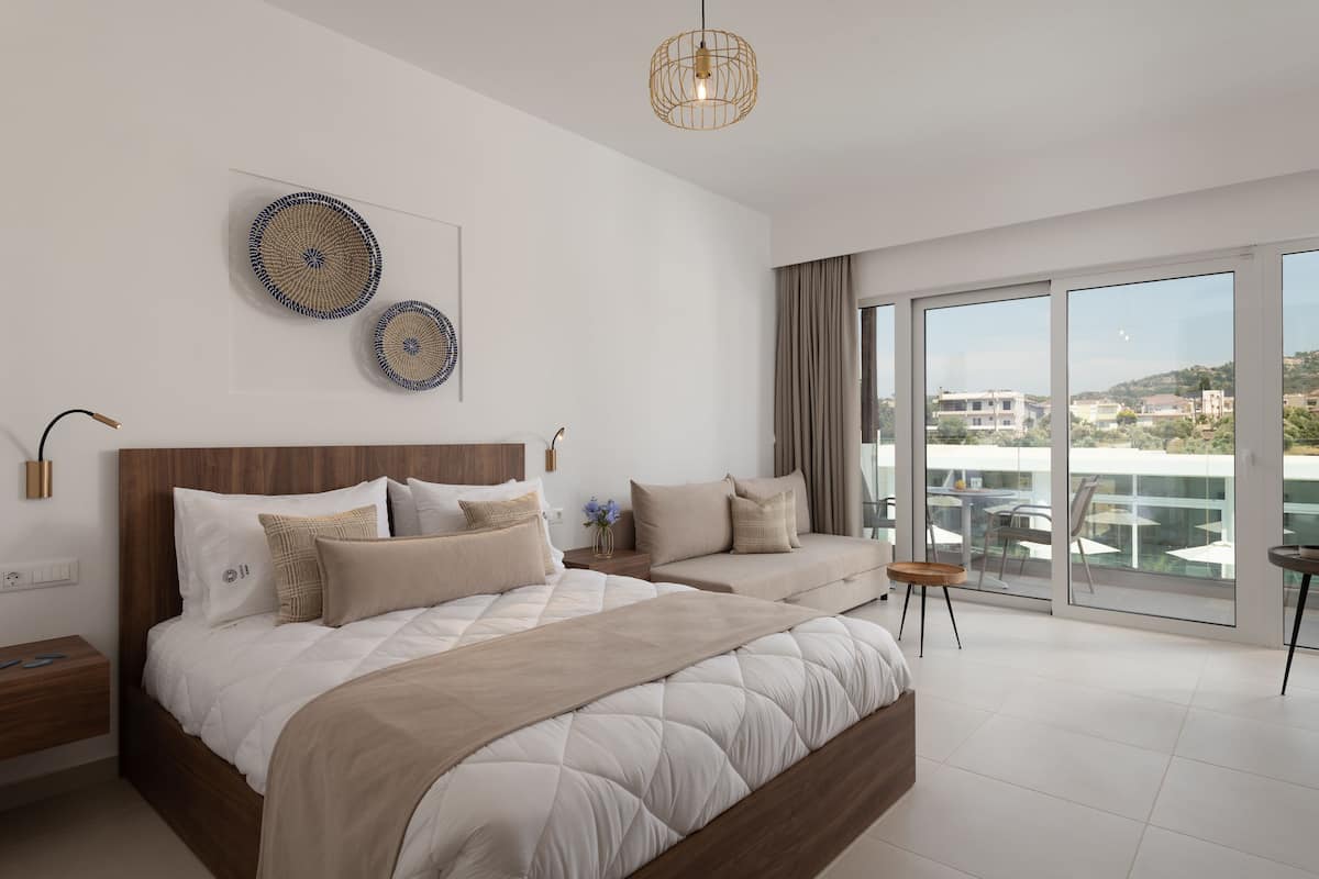 A modern bedroom with a large bed, a sofa, and a balcony with a view of buildings.