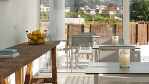 A dining area with a wooden table, chairs, and a view of a windmill and distant buildings through large windows. - Nama Retreat (Rhodes)