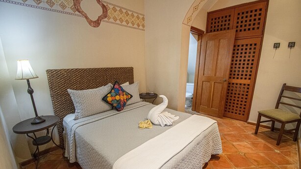 Letto rifatto con copriletto trapuntato, cuscino decorativo e asciugamano piegato. Un comodino con lampada, una sedia e una porta in legno con decoro a griglia.