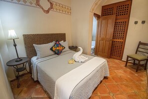 Una cama bien hecha con una colcha acolchada, una almohada decorativa y una toalla doblada sobre la cama. Hay una mesita de noche con una lámpara, una silla y una puerta de madera con diseño de celosía.