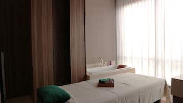 Una habitación de hotel moderna con una cama, un almohadón verde, una mesita de noche, una ventana con cortinas translúcidas y un baño con bañera y lavamanos.