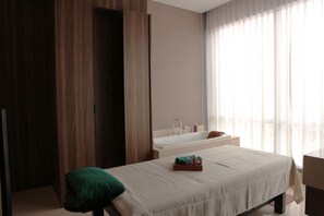 Ein modernes Hotelzimmer mit Bett, grünem Kopfkissen, Nachttisch, Fenster mit durchsichtigen Vorhängen sowie einem Badezimmer mit Badewanne und Waschbecken.