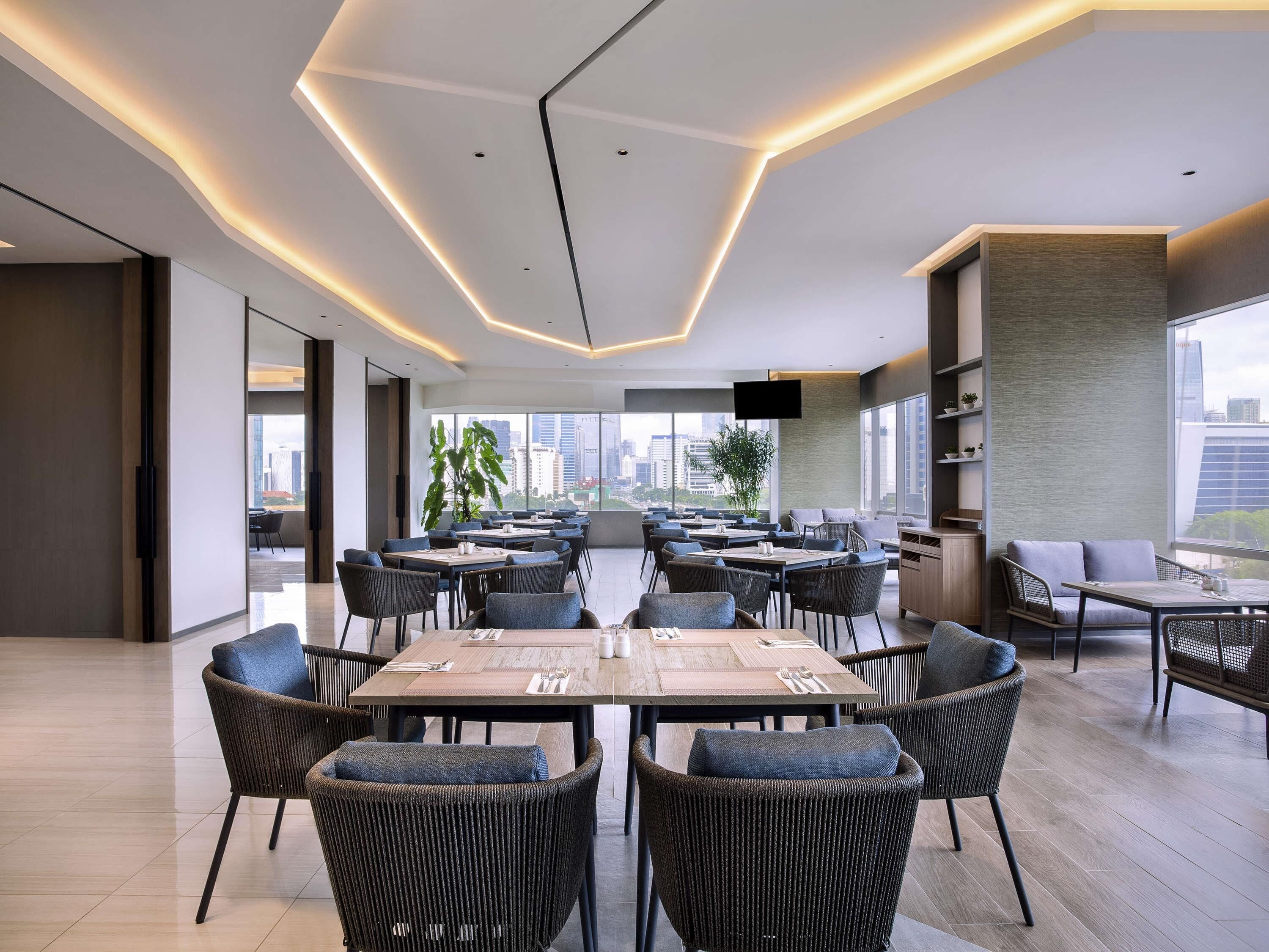 A modern dining area with multiple tables and chairs, large windows offering city views, and a flat-screen TV mounted on the wall.