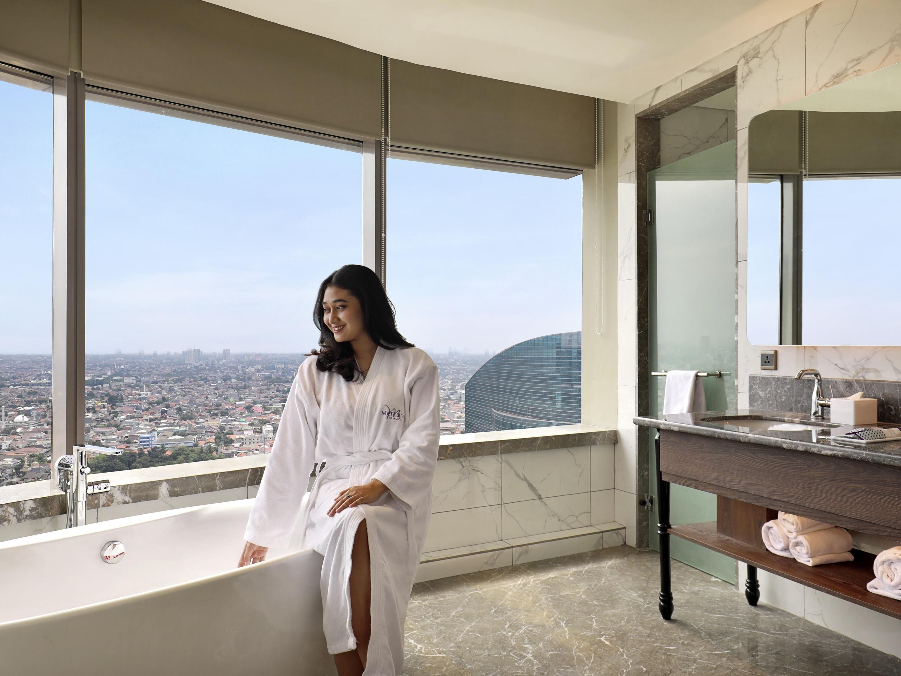 A high-rise hotel room with a large bathtub, a vanity with a mirror, and a city view.