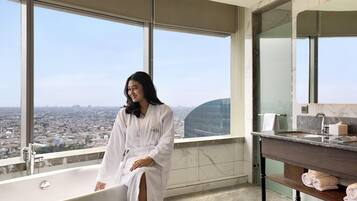 A high-rise hotel room with a large bathtub, a vanity with a mirror, and a city view.