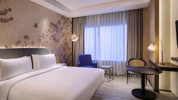 A hotel room with a large bed, a desk, two chairs, a small table, and a large window with curtains.