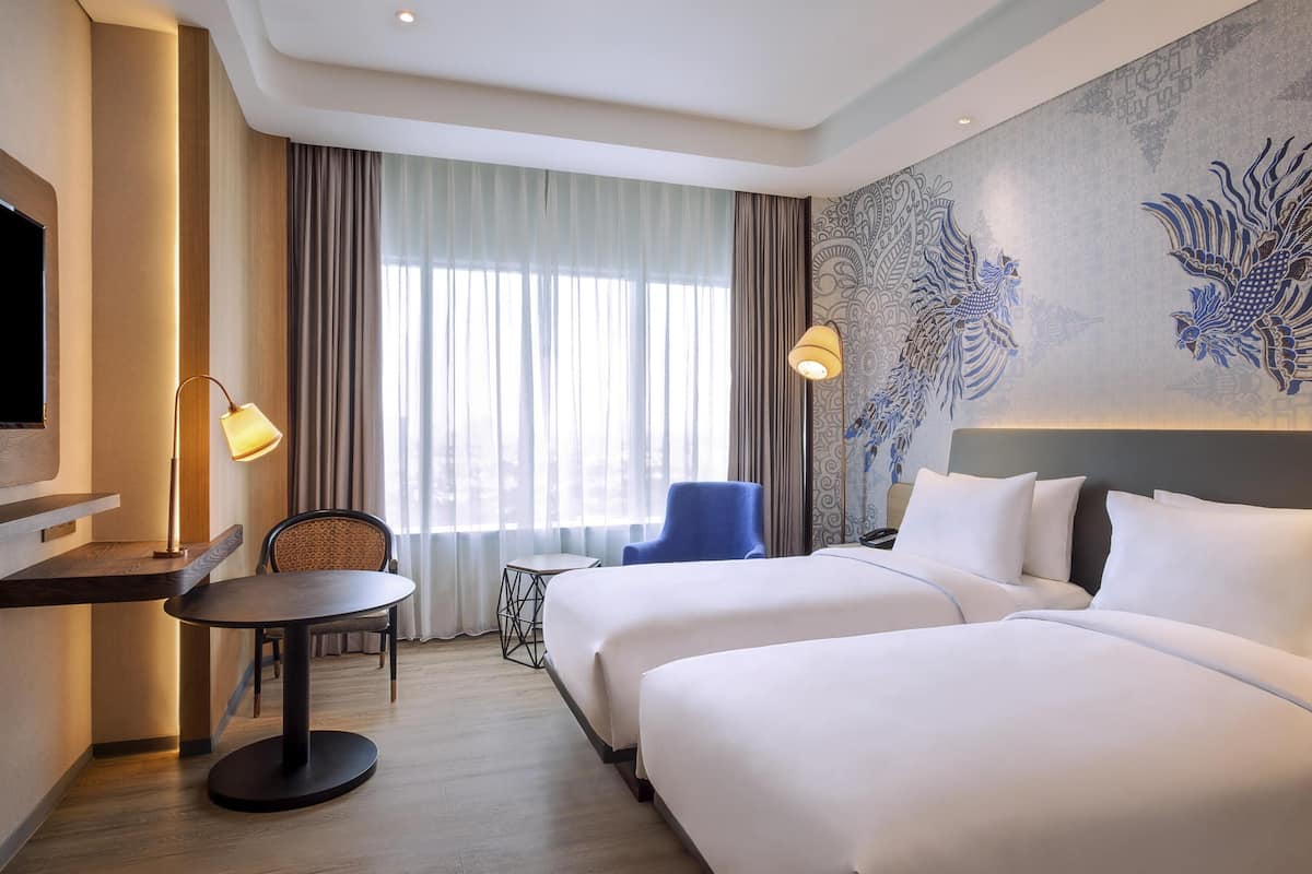 Ein Hotelzimmer mit zwei Betten, einem Schreibtisch, einem Stuhl, einem Fernseher und einem großen Fenster mit Vorhängen.