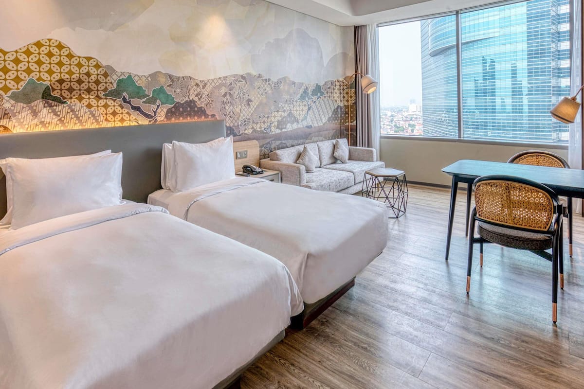 Ein Hotelzimmer mit zwei Betten, einem Schlafsofa, einem Esstisch und einem großen Fenster mit Blick auf die Stadt.