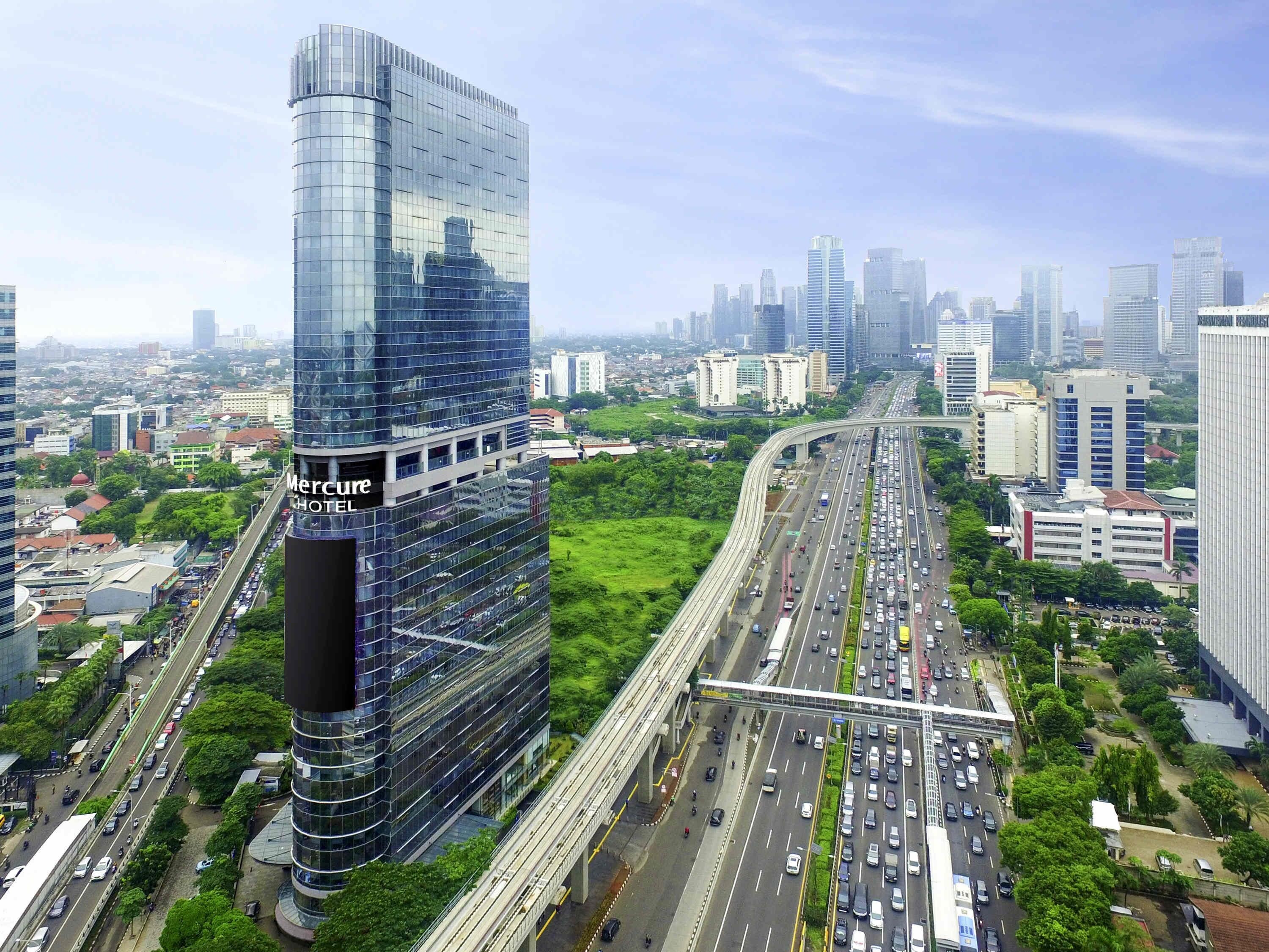 Un moderno hotel de gran altura con fachada de vidrio curvada, situado cerca de una autopista con mucho tráfico y rodeado de edificios urbanos.