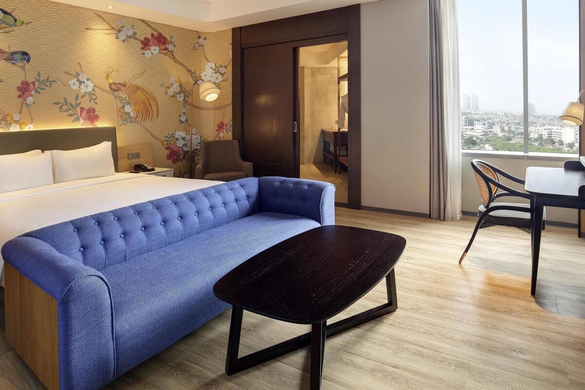 Ett hotellrum med en säng, en blå soffa, ett litet bord, en stol, en vägg med blommigt tapetmönster och ett stort fönster med utsikt över staden.