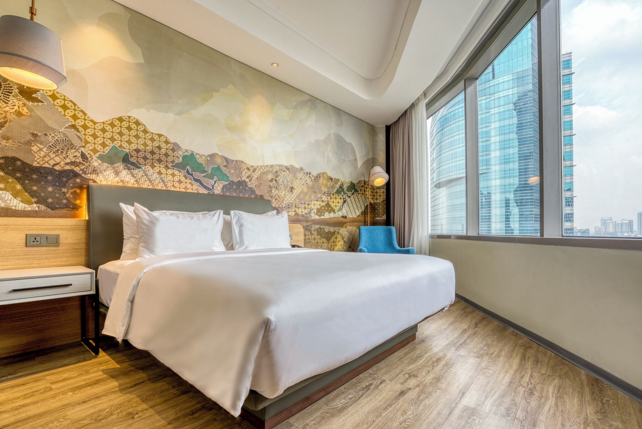 A modern hotel room with a large bed, a nightstand, a lamp, and a view of the city through large windows.