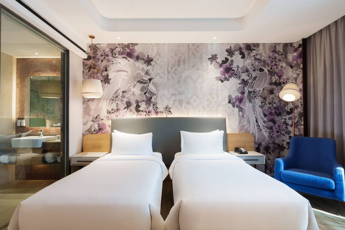 Ett hotellrum med två sängar, en blå fåtölj, en stor väggmålning med blommotiv och ett modernt badrum som syns genom en glasvägg.