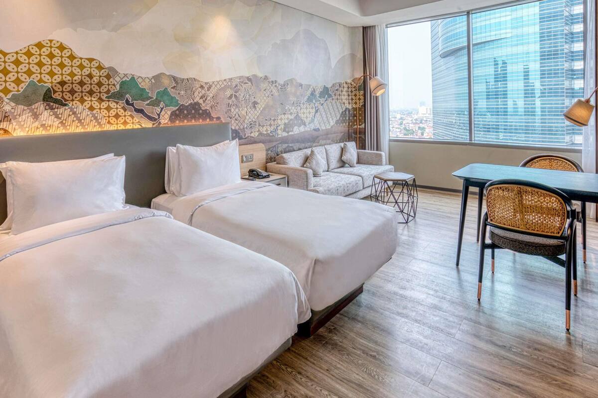 Ett hotellrum med två sängar, en soffa, ett matbord och ett stort fönster med utsikt över staden.