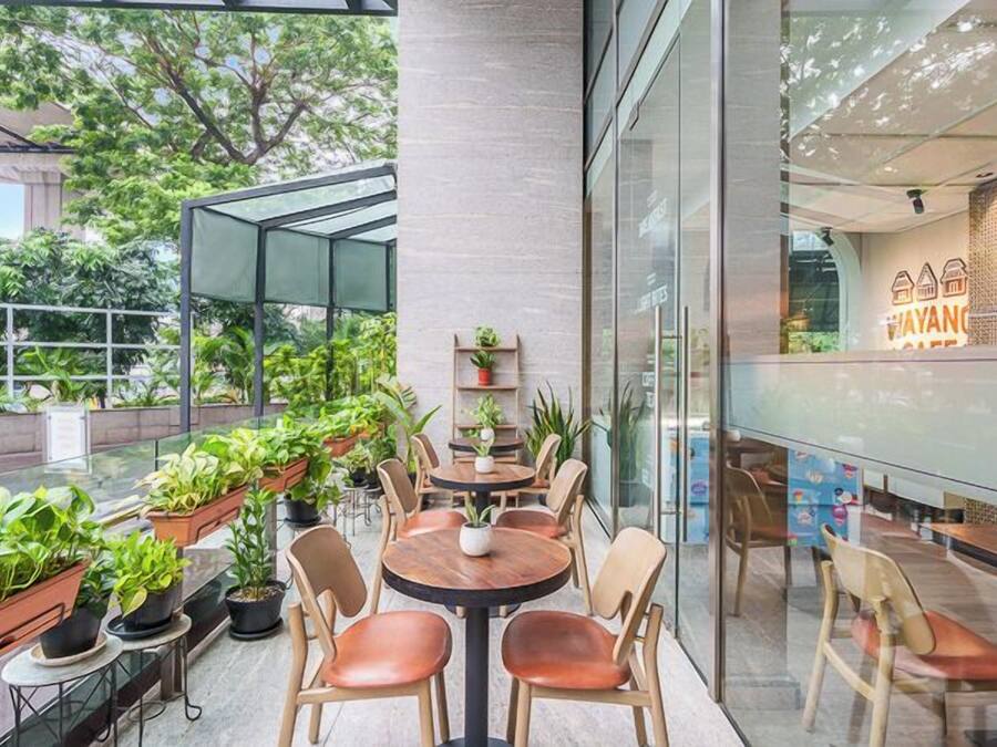 Un área de estar moderna al aire libre con mesas y sillas redondas, rodeada de plantas en macetas y paredes de vidrio.