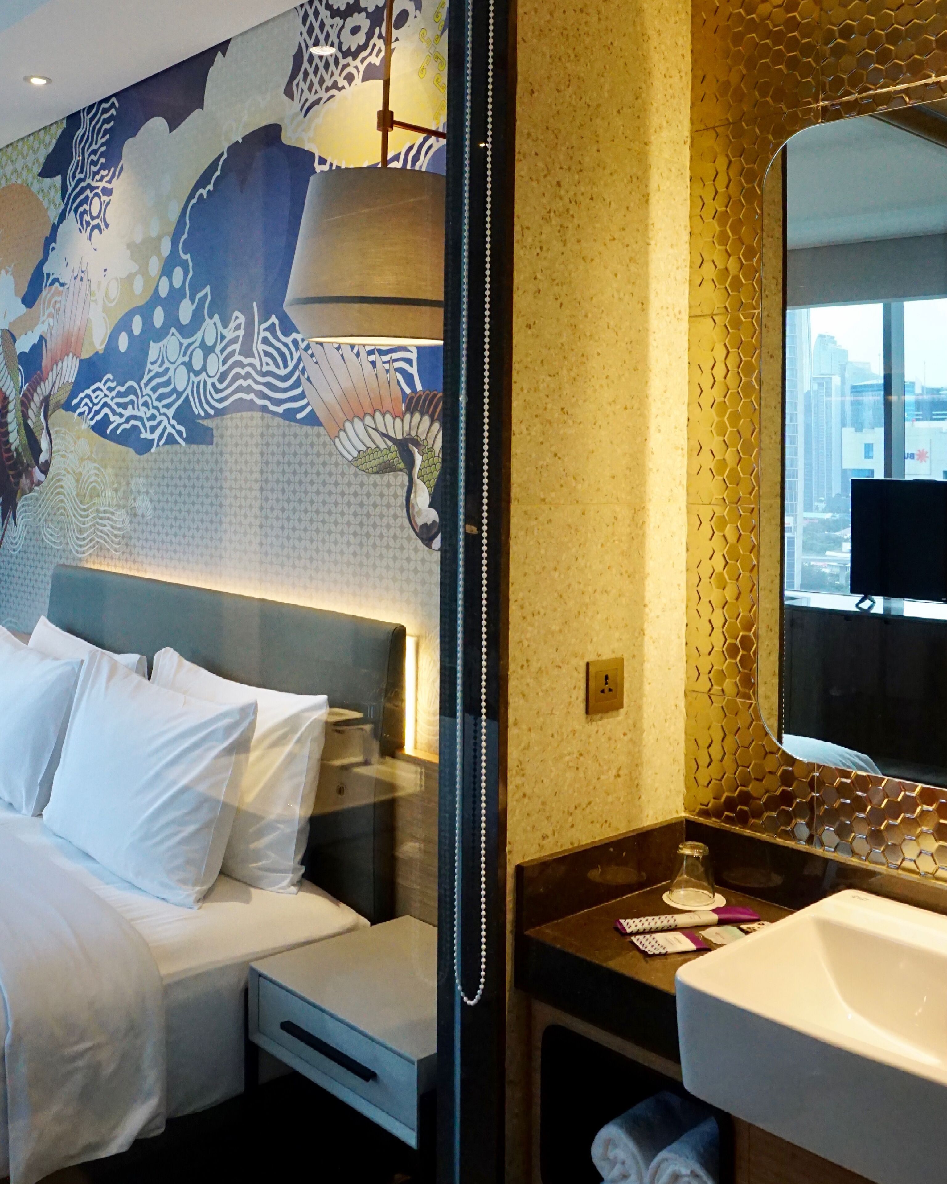Habitación de hotel con cama, espejo grande, lavamanos y vistas a la ciudad.